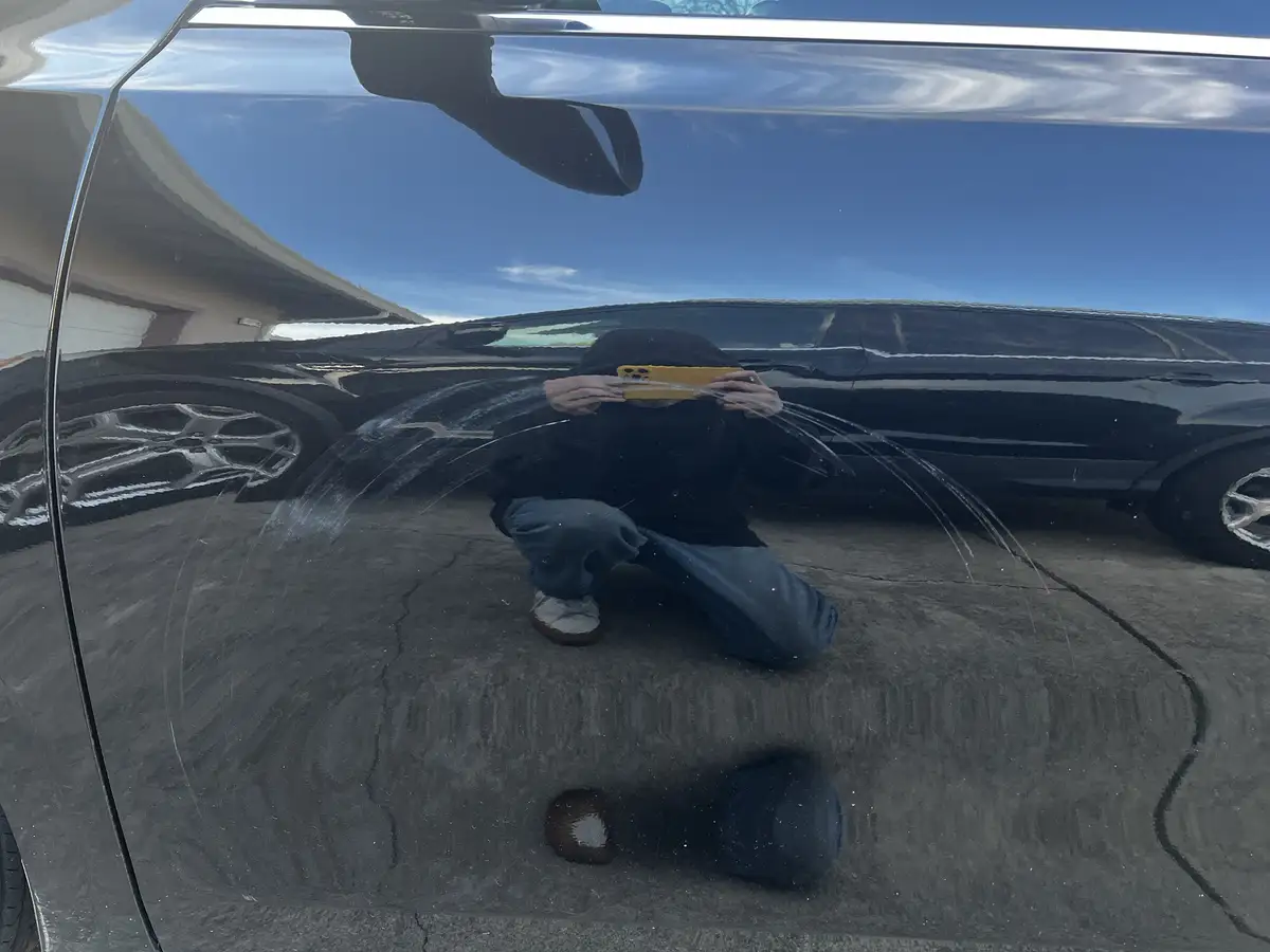 A person crouching on a driveway is reflected in the glossy black surface of a car, holding a phone with a yellow case to…
