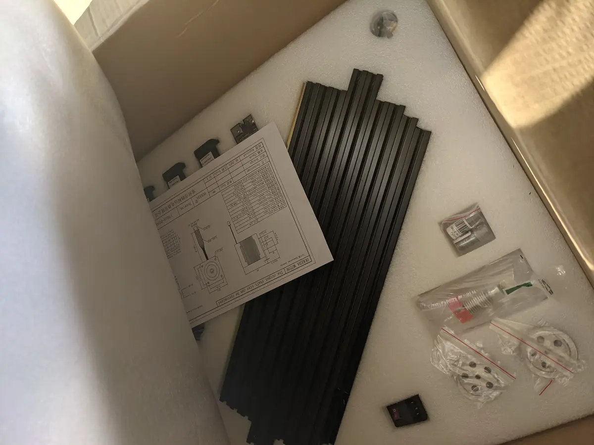 An overhead shot of an open cardboard box filled with white foam padding and various unassembled mechanical parts, including…