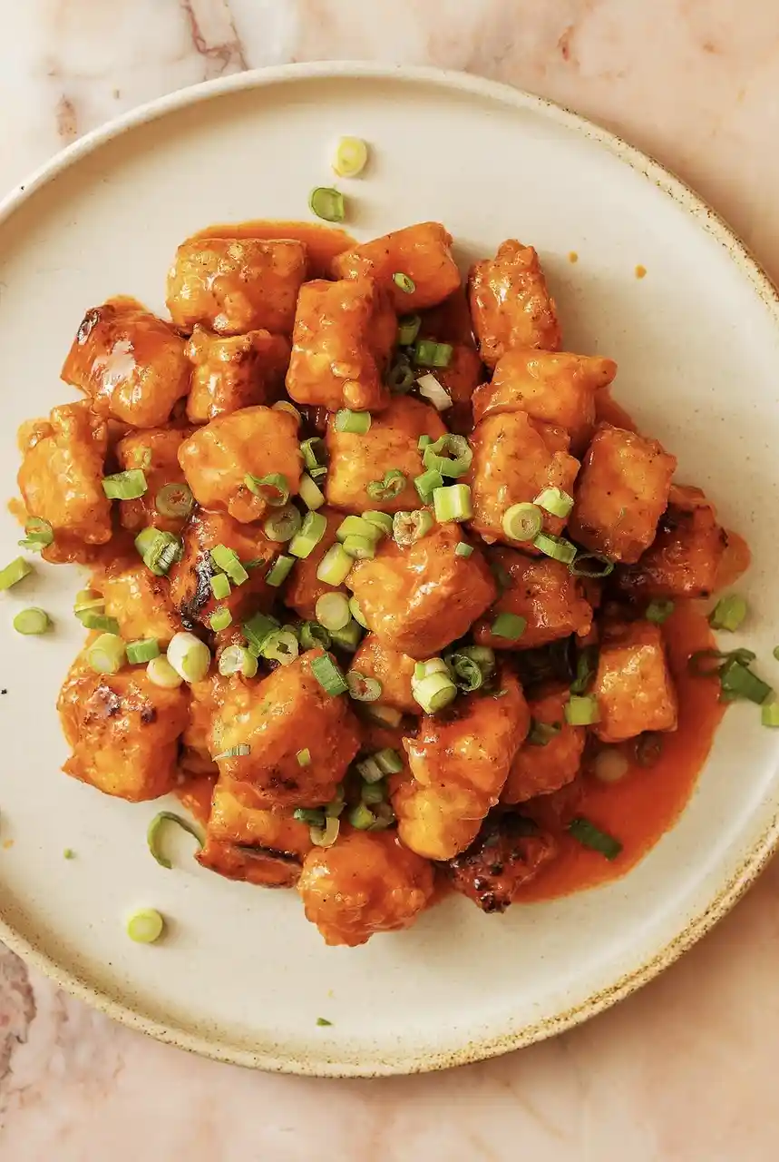 A top-down photo of a plate of orange-brown glazed chicken pieces topped with chopped green onions, set on a light-colored…