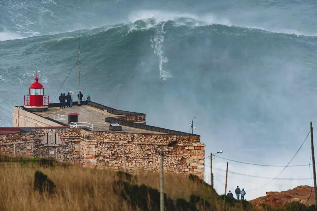 A surfer rides a massive, towering wave while a red lighthouse and stone fortification stand on the shore. Several people…