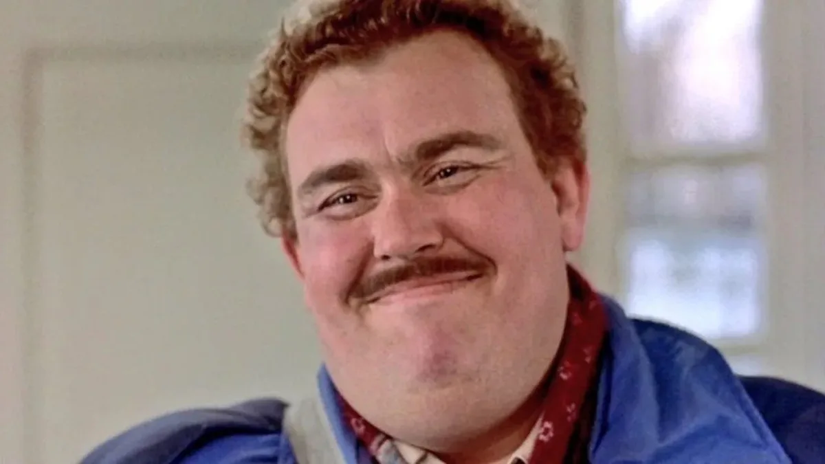 a close-up of a man with curly reddish hair, a mustache, and a smug, self-satisfied grin, wearing a blue jacket over a red…