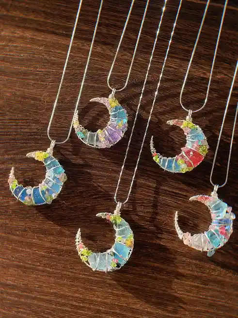 Five handmade crescent moon necklaces, each crafted with wire-wrapped colorful gemstones, are displayed on a dark wooden…