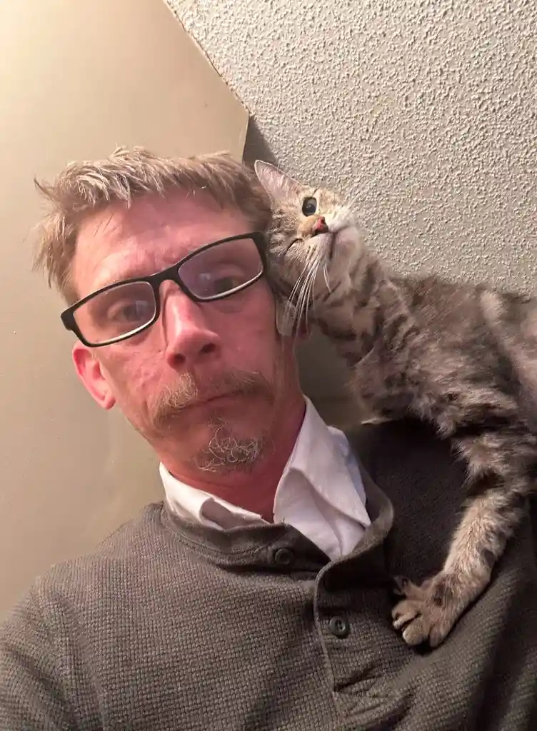 A man with glasses and a mustache looks at the camera while a tabby cat rests on his shoulder, looking upward. The text…