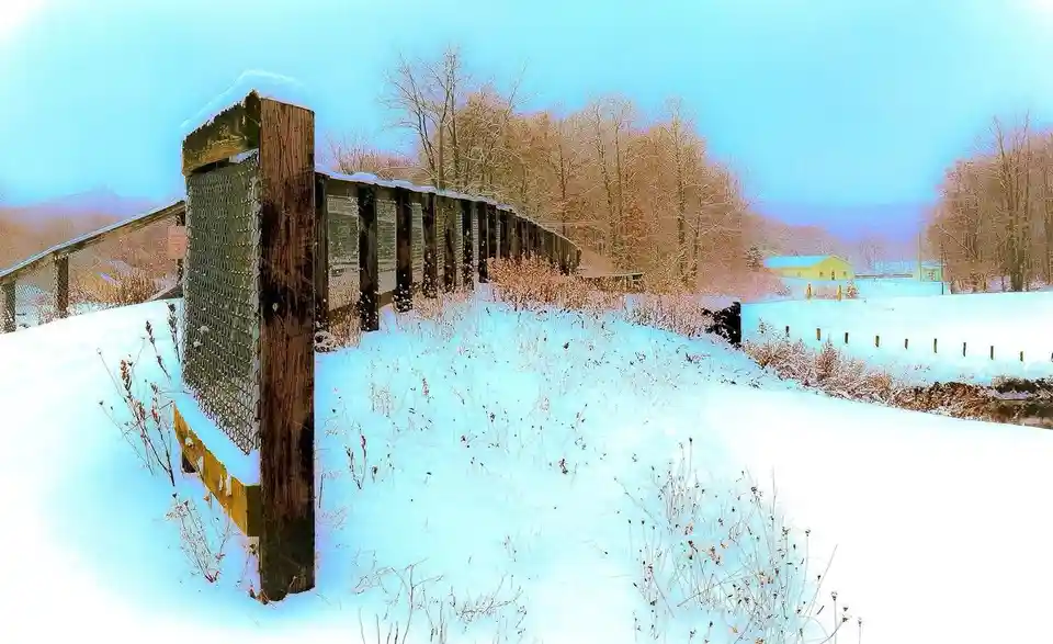 A snowy rural landscape features a wooden fence post with chain-link fencing, partially buried in snow, with bare trees and…