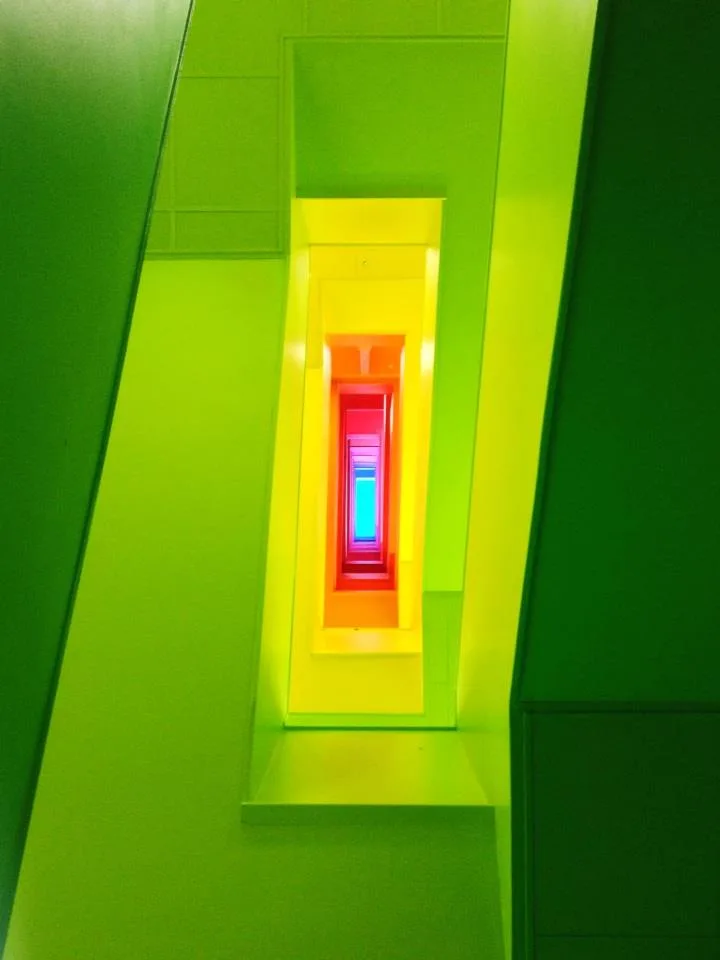 A photograph of a brightly lit, receding hallway with walls in progressively changing neon colors, starting with green on…