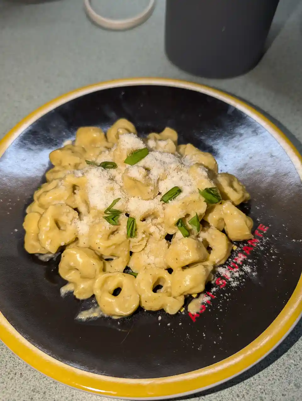 A close-up photo shows a black plate with a yellow rim, holding a serving of tortellini in a creamy sauce, topped with…
