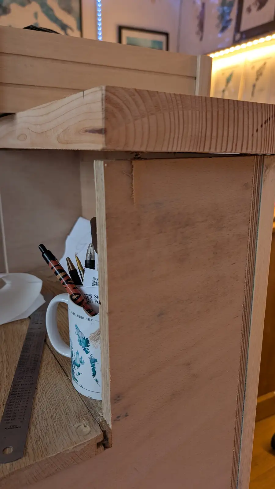 A close-up shot of a wooden desk or shelf corner, with a white mug holding pens and pencils peeking out from behind a wooden…