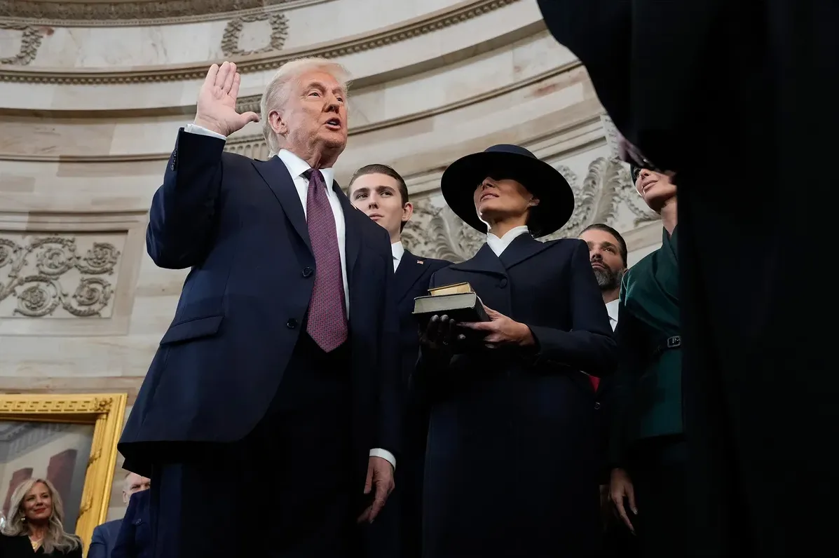 a photo of Donald Trump raising his right hand while taking the presidential oath of office, with Melania Trump holding a…