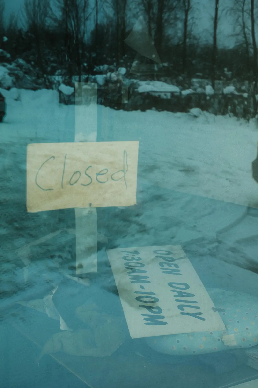 A close-up of a glass storefront window shows two handwritten signs taped to it. The top sign, on beige paper, reads…
