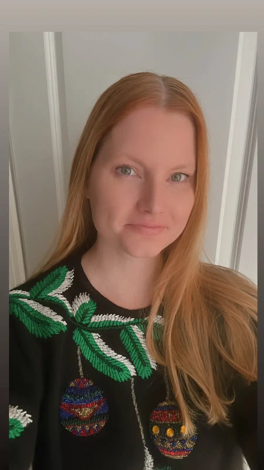 A woman with long red hair and a nose ring looks at the camera with a slight smirk while wearing a black sweater decorated…
