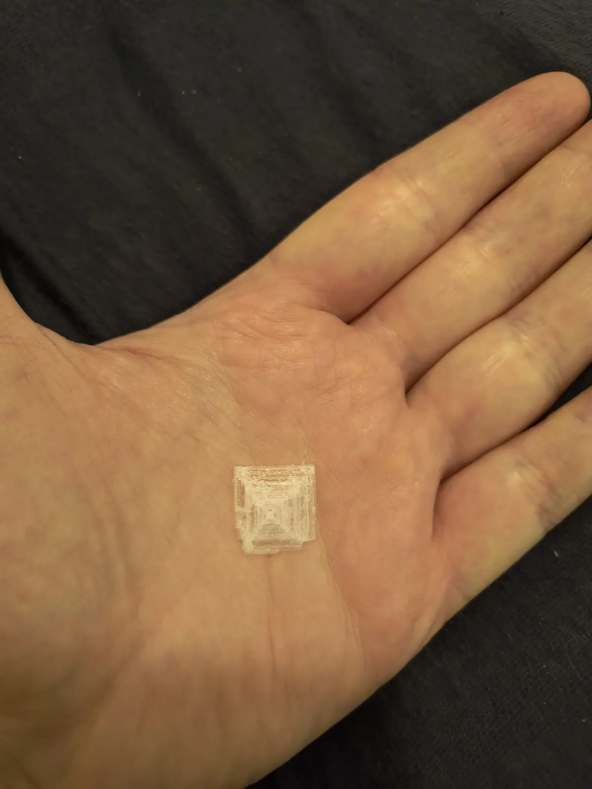A close-up photo shows a person's open palm holding a small, translucent, 3D-printed cube with visible layer lines. The cube…