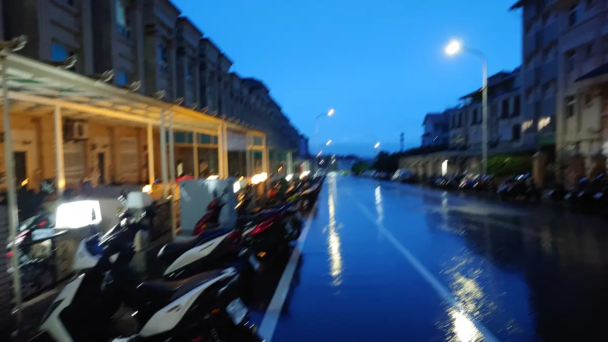 A blurry, motion-blurred photo of a wet city street at dusk, lined with parked scooters on both sides, streetlights…
