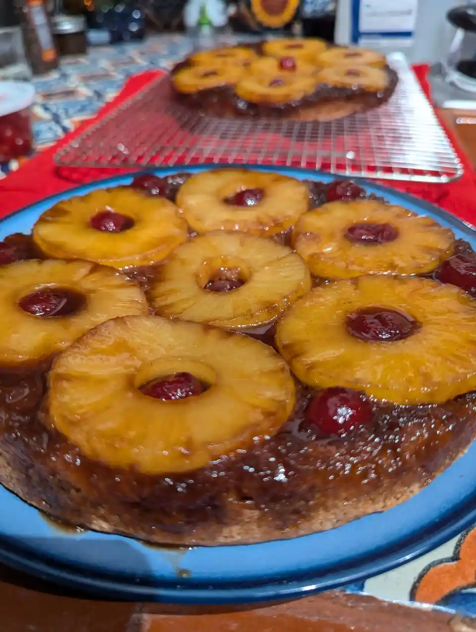 A close-up photo shows a freshly baked pineapple upside-down cake on a blue plate, topped with golden pineapple rings and…