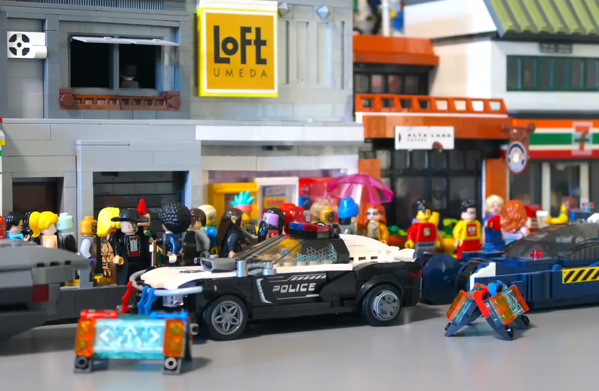 A detailed Lego diorama depicts a bustling city street scene with a black and white police car parked in front of a building…