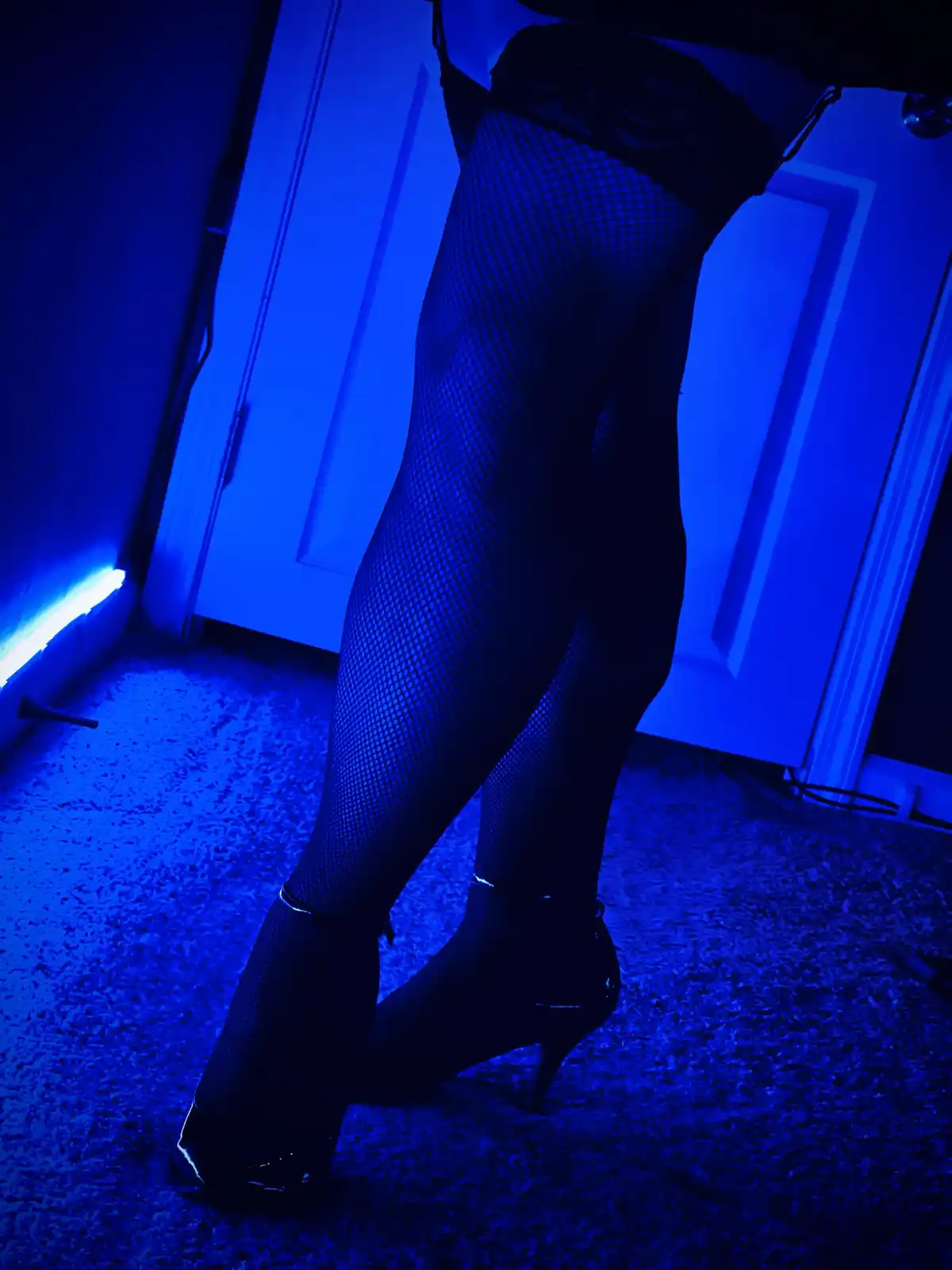 A low-angle shot shows a person's legs clad in black fishnet stockings and high-heeled shoes, standing on a carpeted floor…