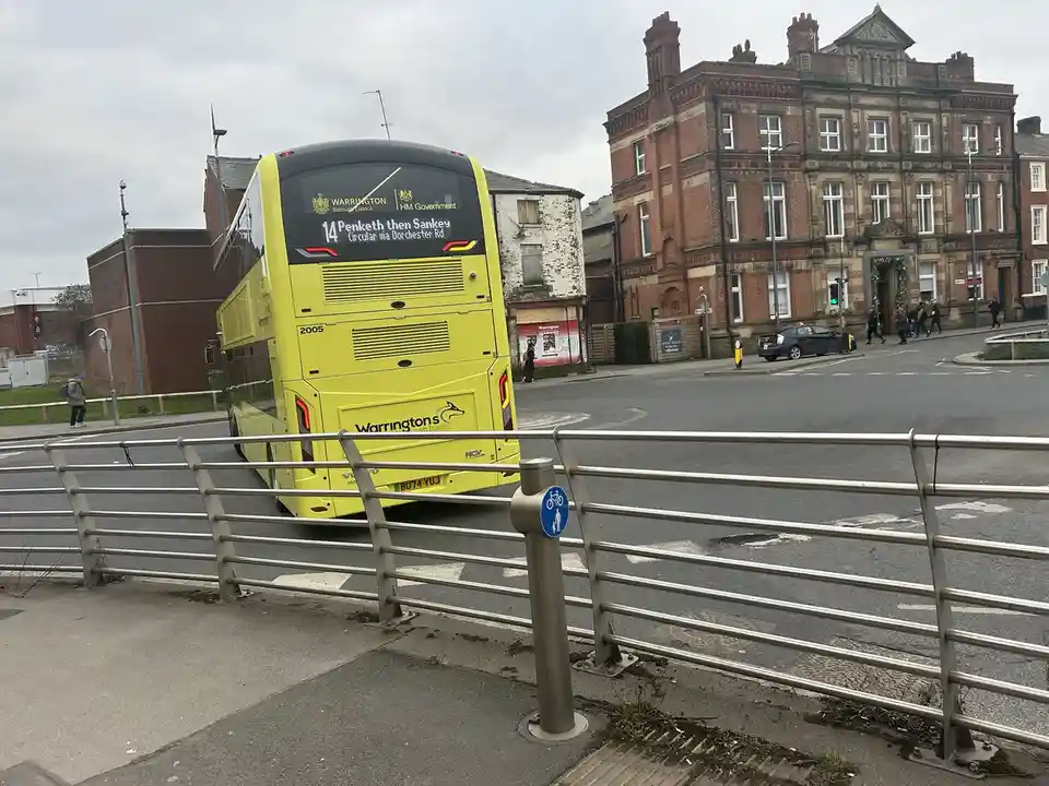 A yellow double-decker bus with route 14 to Penketh then Sankey is seen from behind, stopped at a street corner in…