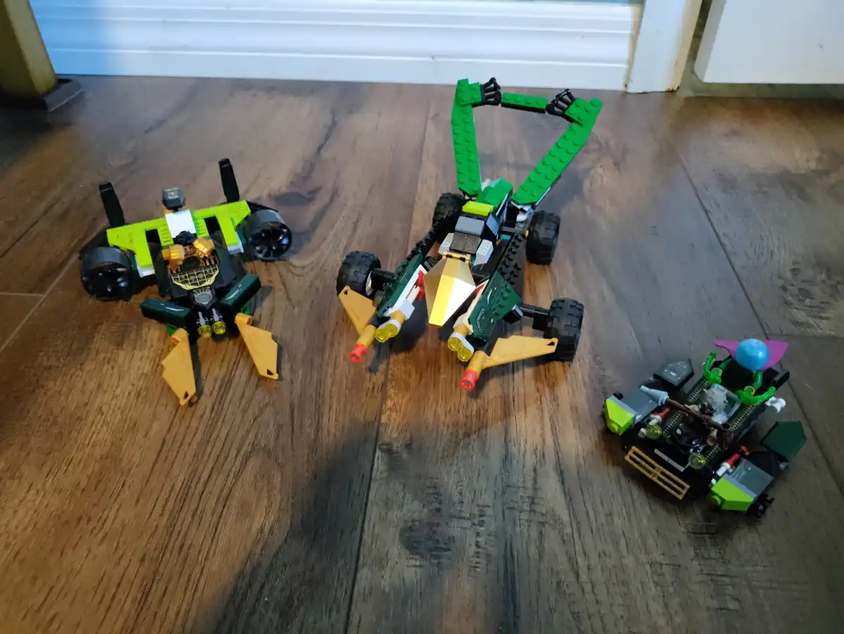 Three LEGO Ninjago vehicles are arranged on a wooden floor, with the central one having large green wings raised, resembling…