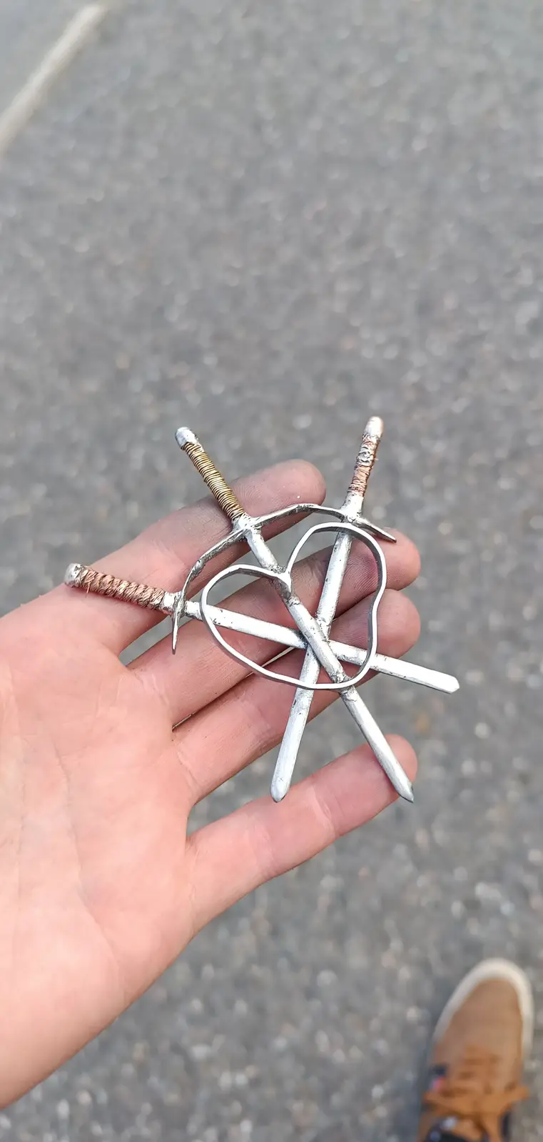 A hand holds a small, handmade metal object resembling a sword with a heart-shaped guard, set against an asphalt background…