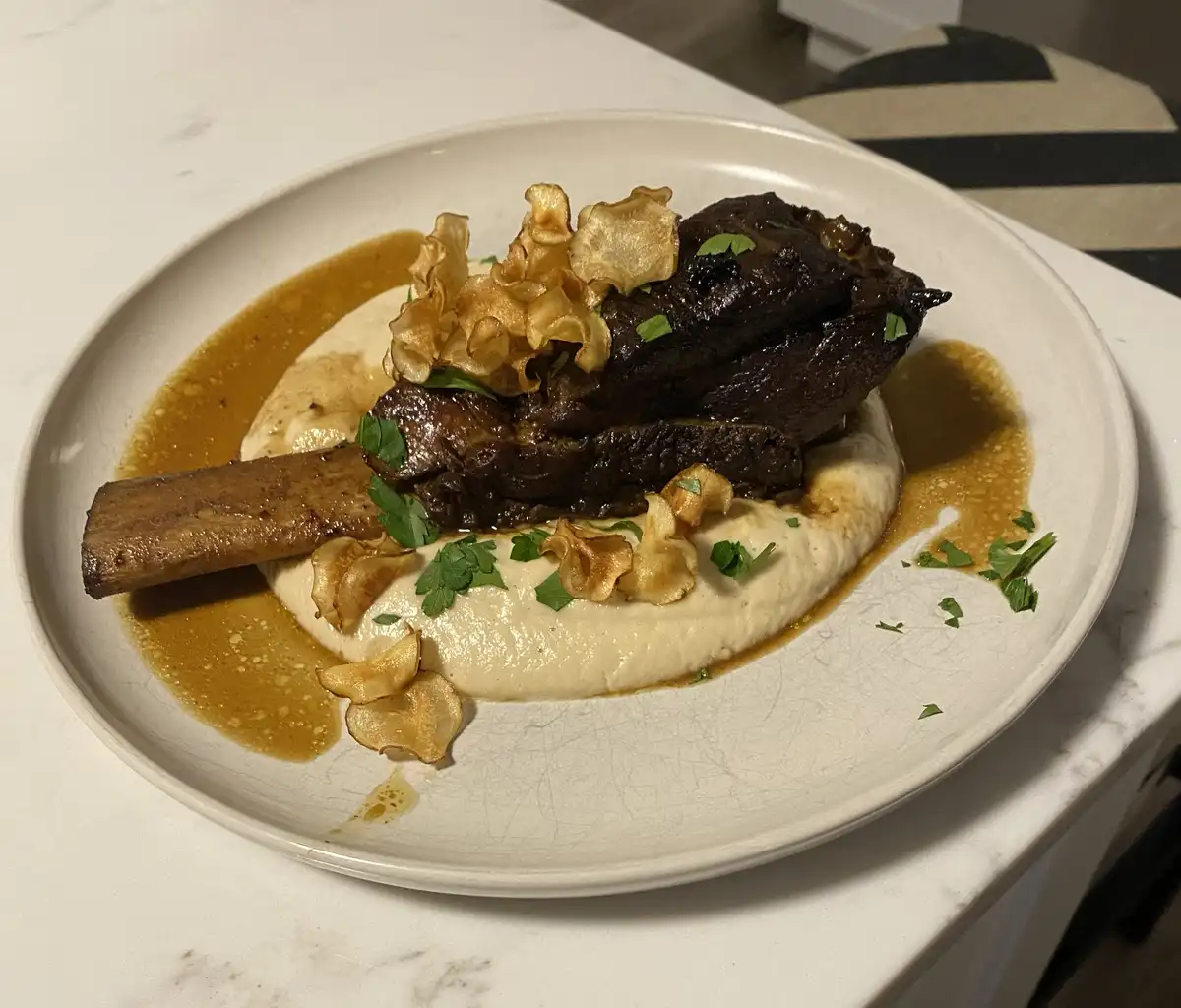 A close-up photo of a plated dish featuring a large, dark, bone-in meat piece resting on a creamy white puree, all drizzled…