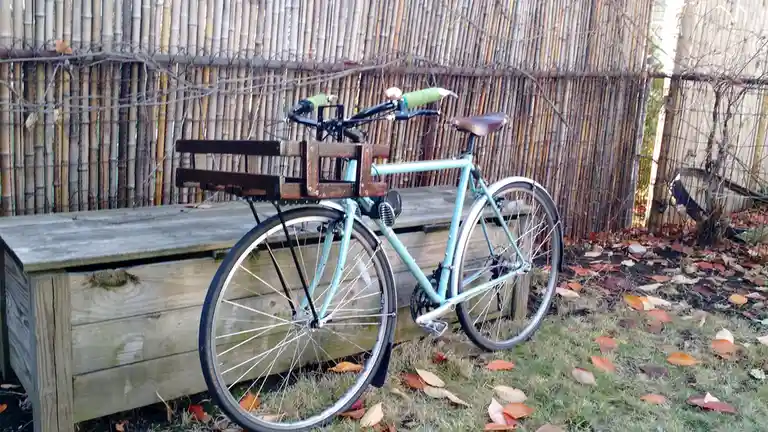 A light blue bicycle with a wooden basket is parked outdoors on grass scattered with autumn leaves, leaning against a wooden…