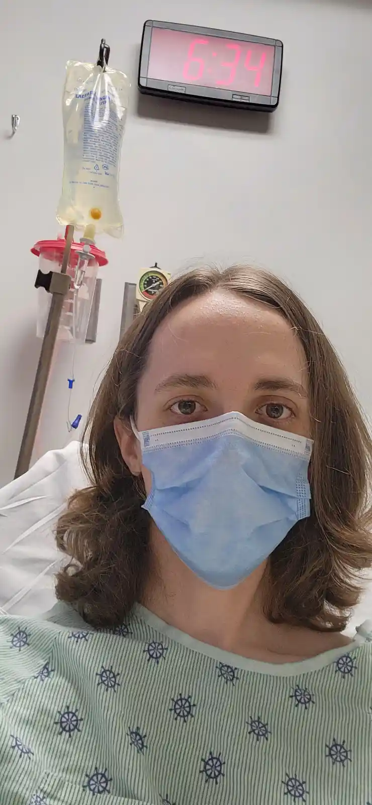 A person wearing a blue surgical mask and a green hospital gown with ship wheel patterns looks at the camera from a hospital…