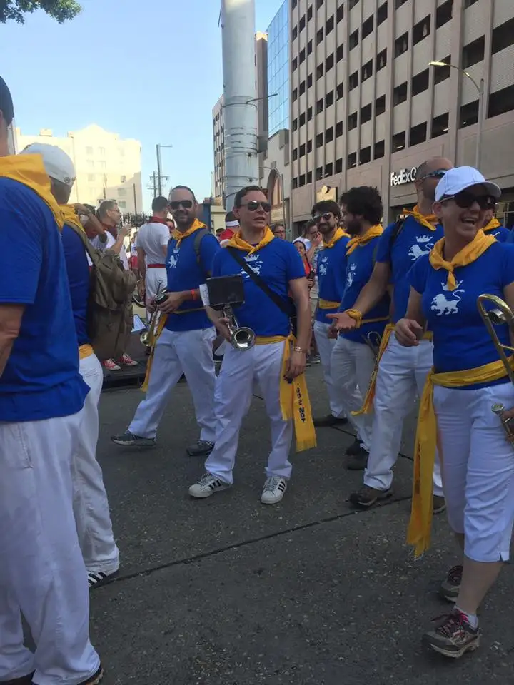A group of people in matching blue shirts and white pants, with yellow scarves around their necks, are marching in a street…