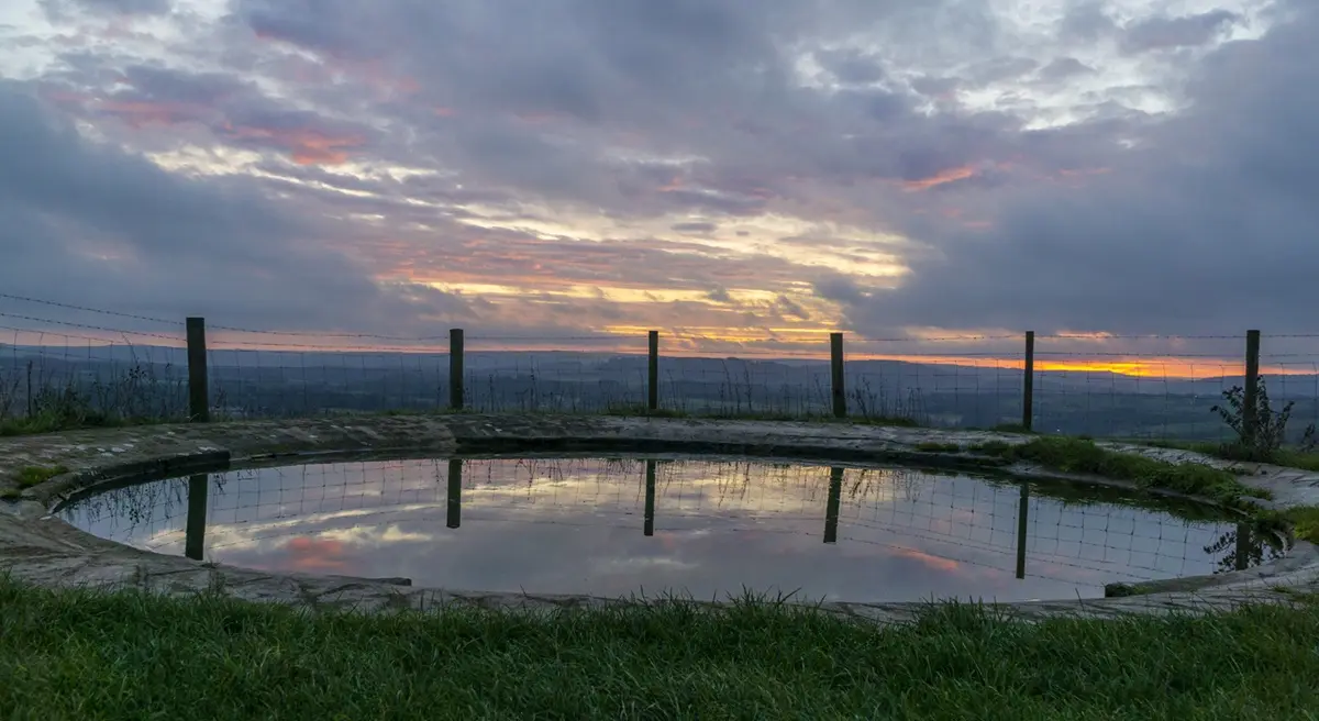 A serene, circular stone-edged pond reflects a dramatic sunset sky with pink, orange, and purple clouds. A barbed wire fence…