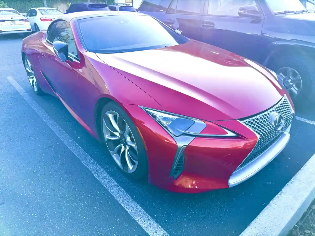 A shiny red Lexus LC 500h sports car is parked in a lot, its sleek design and distinctive grille visible under bright…