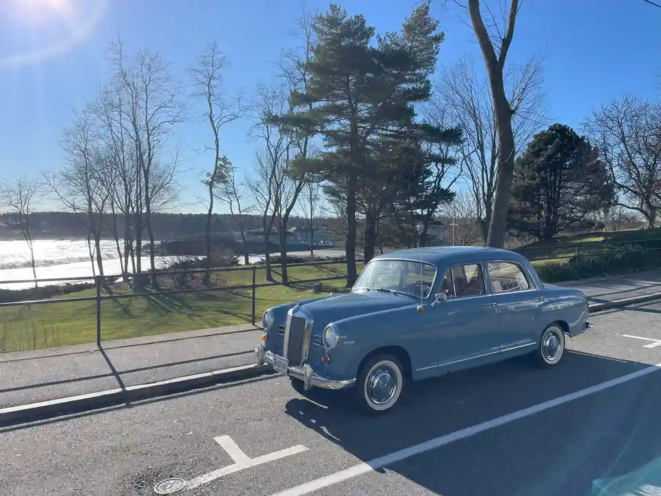 A vintage blue Mercedes-Benz sedan is parked in a sunny parking lot next to a grassy area with trees and a body of water in…