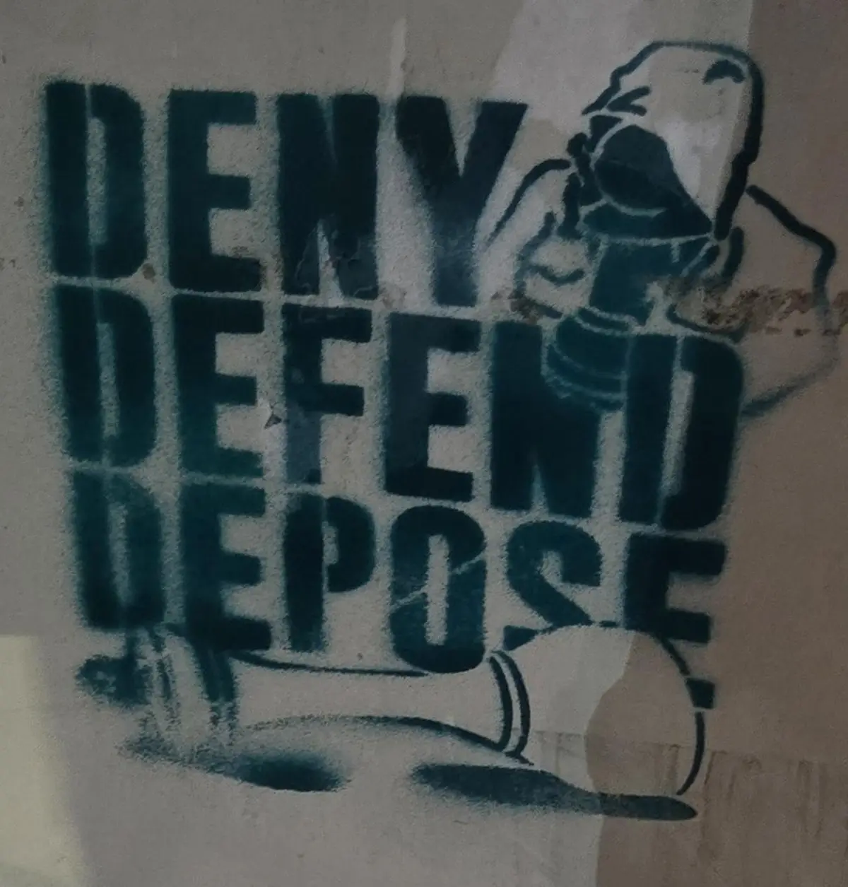 A stencil graffiti on a wall shows the words 'DENY', 'DEFEND', and 'DEPOSE' stacked vertically in bold, teal letters. To the…