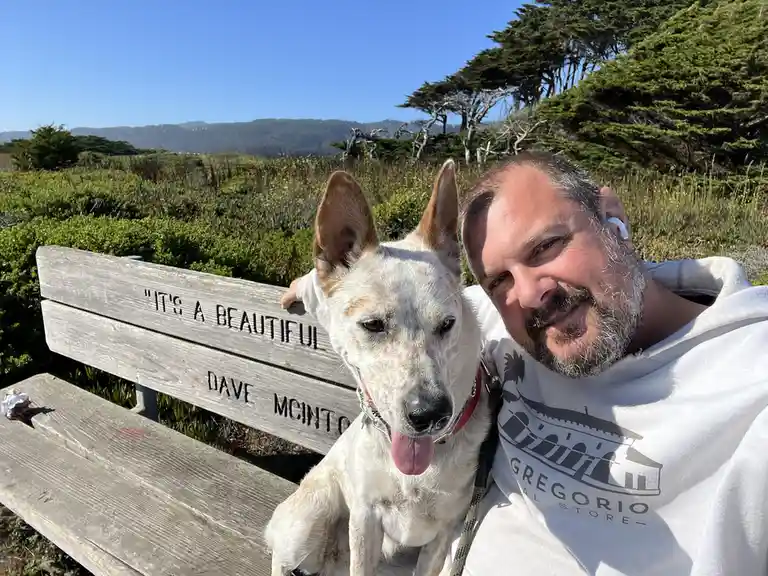 A man with a beard and white earbuds sits on a wooden bench outdoors with a white dog that has its tongue out, looking…