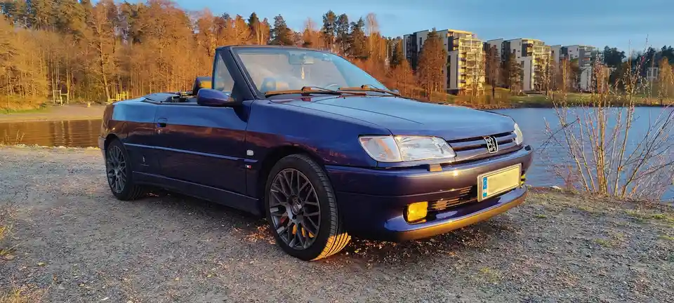 A dark blue Peugeot 405 convertible is parked on a gravel shore beside a calm lake, with bare trees and modern apartment…