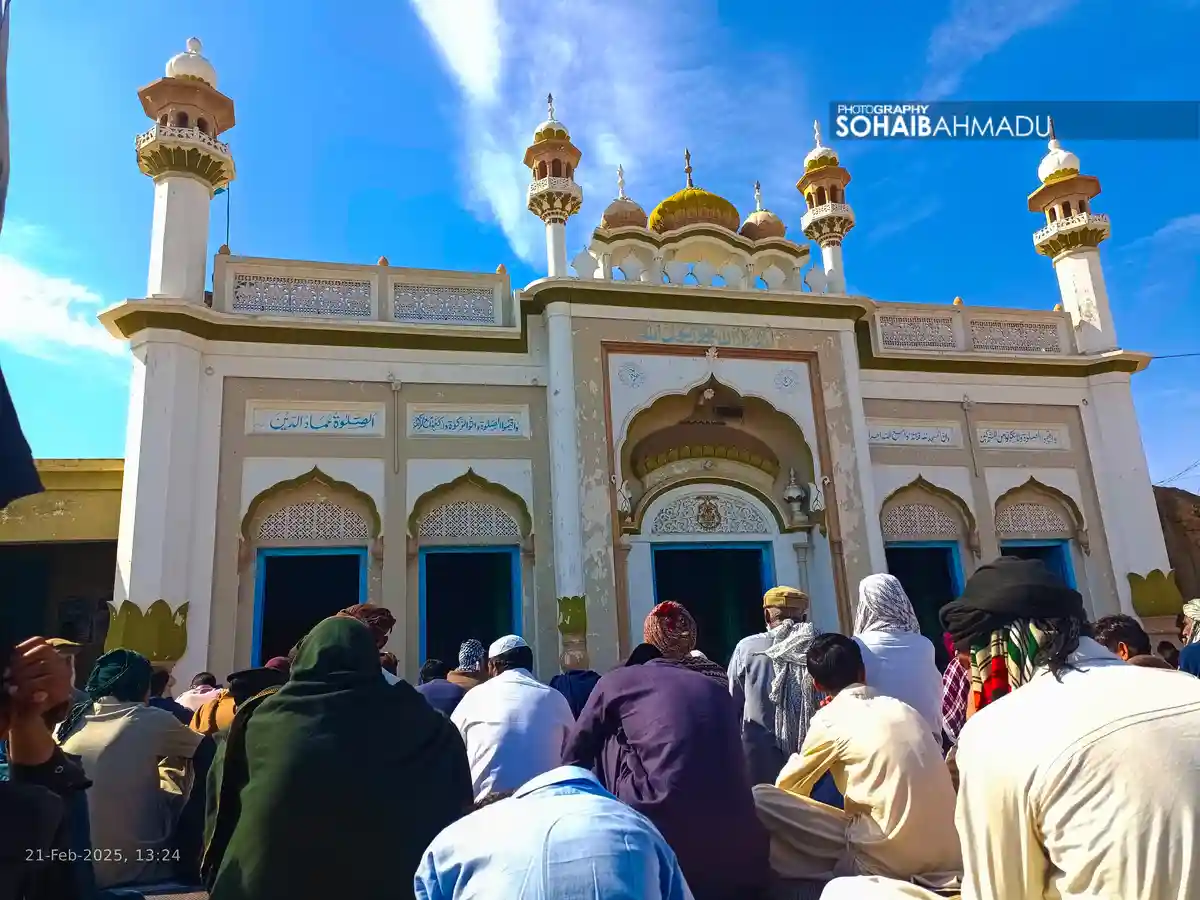 A group of people are seen from behind, sitting and praying in front of a white mosque with blue doors, golden domes, and…