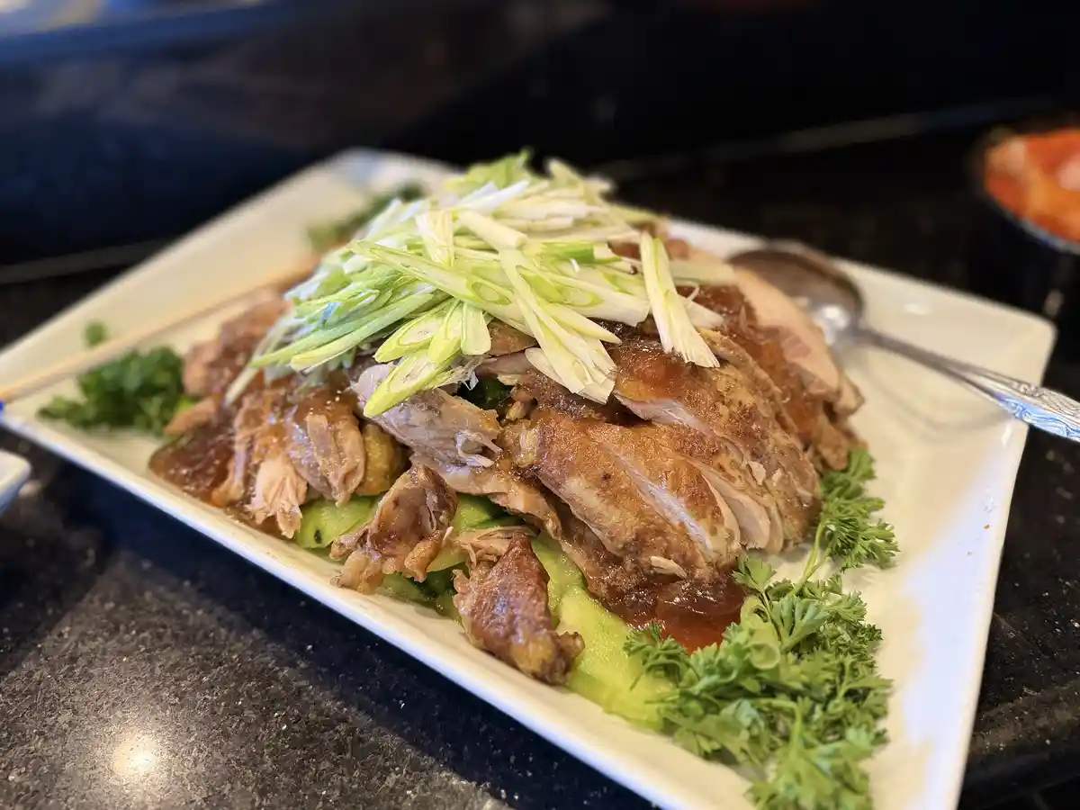 A close-up photo of a plate of sliced roasted duck with a glossy sauce, topped with chopped green onions and garnished with…