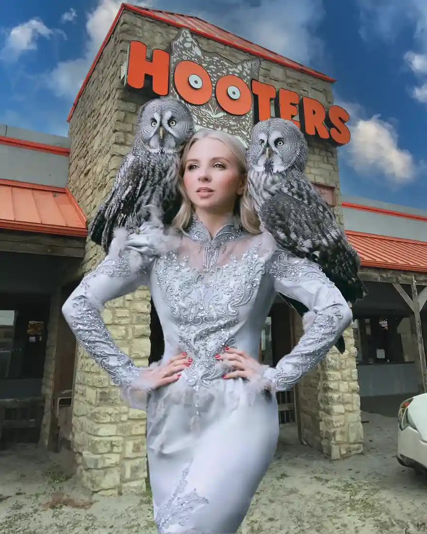 A woman in an ornate silver dress stands confidently with two large owls perched on her shoulders, in front of a Hooters…