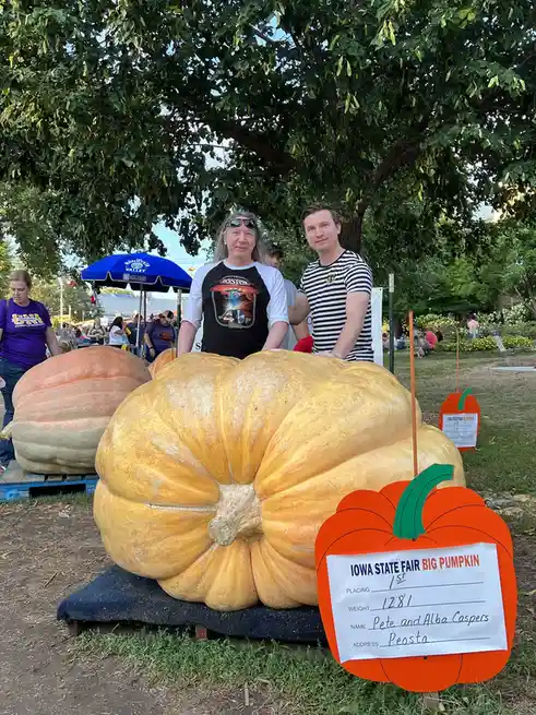 Two men stand proudly behind a massive, yellow-orange pumpkin at the Iowa State Fair, with a sign in front reading 'IOWA…