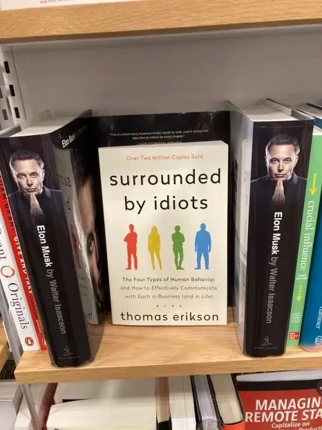 A bookshelf displays a book titled 'surrounded by idiots' by Thomas Erikson, flanked on both sides by books featuring Elon…