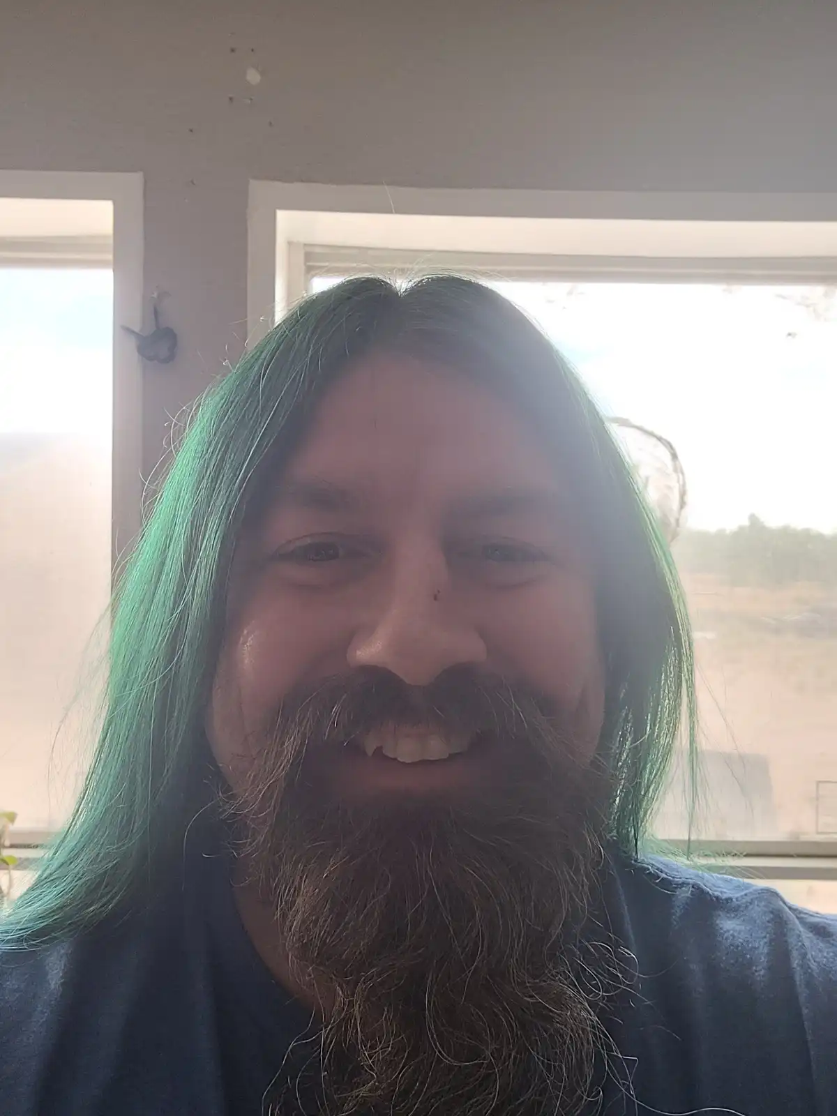 A man with long, vibrant green hair and a full beard smiles at the camera while standing indoors near windows. The overlaid…