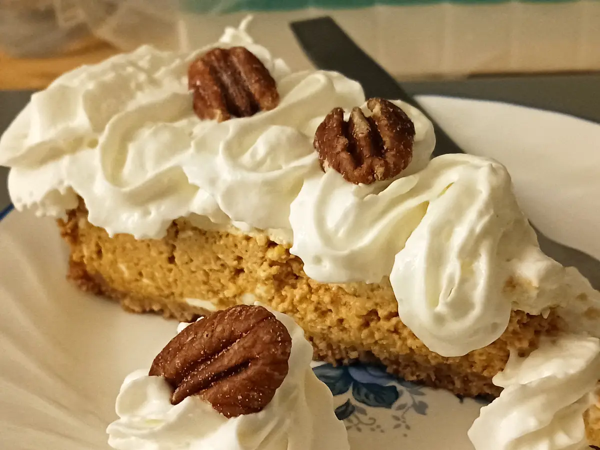 A close-up photo shows a slice of pumpkin pie with whipped cream and pecans on top, served on a white plate with a blue…