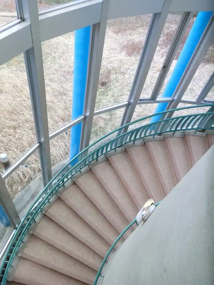 An overhead view of a spiral staircase with a teal railing and beige steps, surrounded by large glass windows revealing dry…