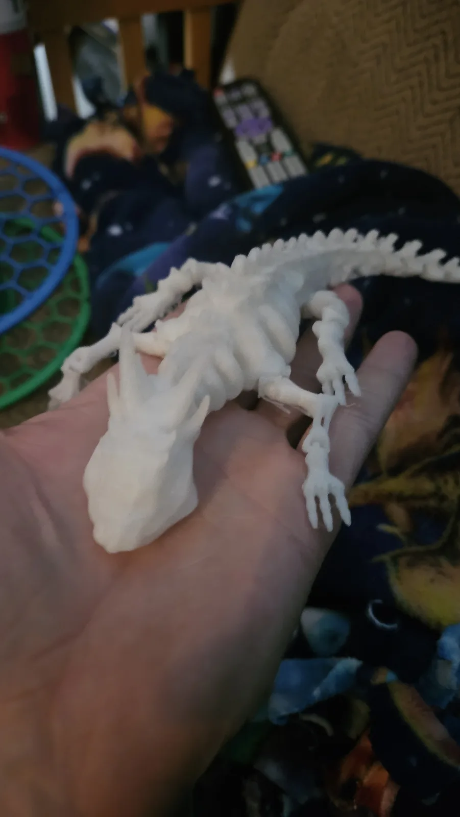 A person holds a small, white 3d-printed lizard skeleton in their open palm. The background is a blurry indoor scene with a…