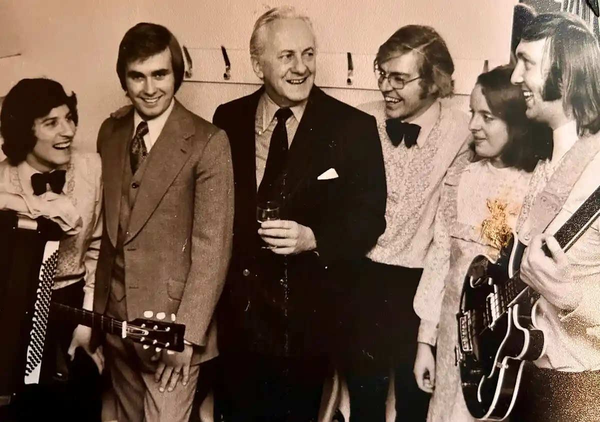 A sepia-toned vintage photo shows six people, likely a band, smiling and posing together in 1970s attire. Several hold…