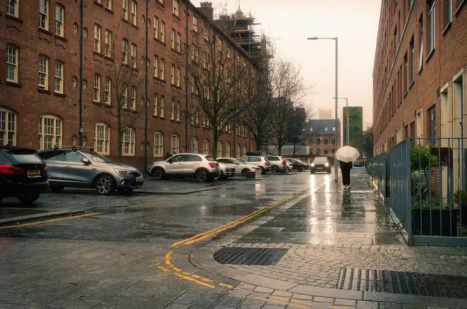 A person walks down a wet, rainy urban street holding a white umbrella with black polka dots, away from the camera. Cars are…
