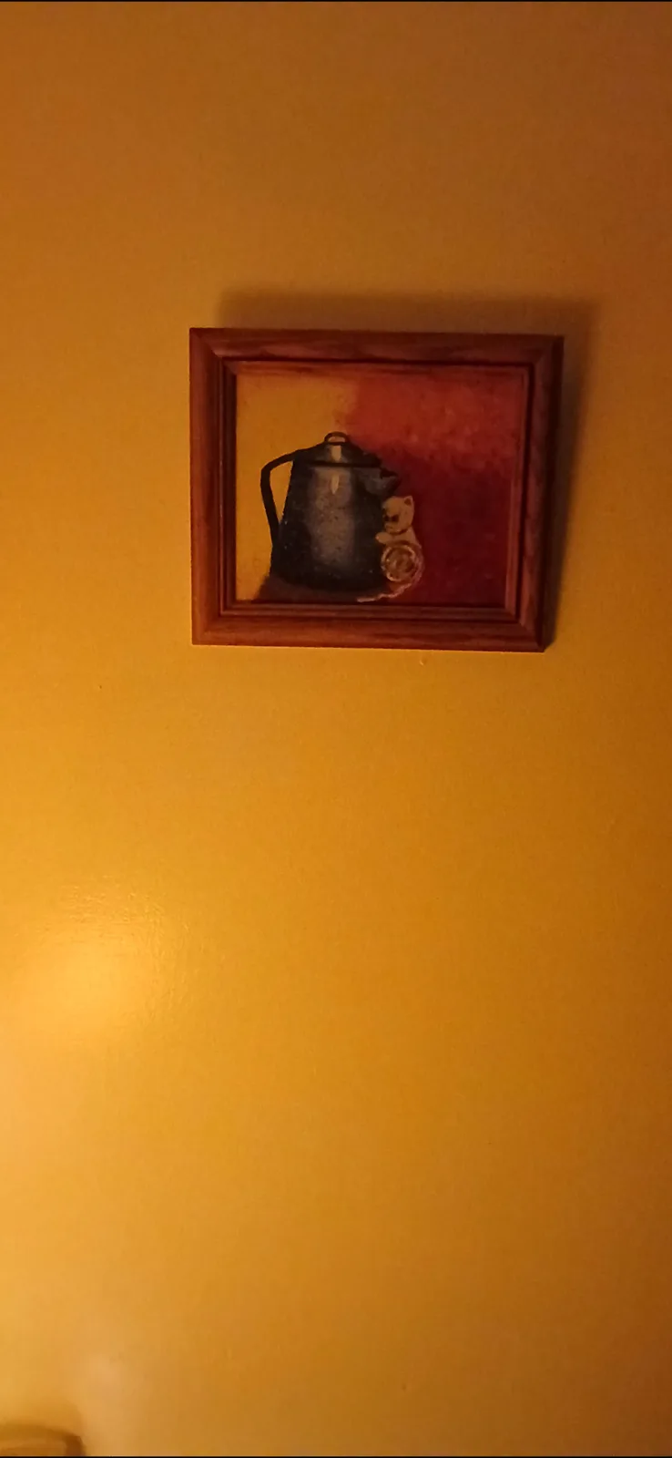 A framed painting hangs on a warm orange wall, depicting a dark metal kettle with a small cat curled up beside it, seemingly…