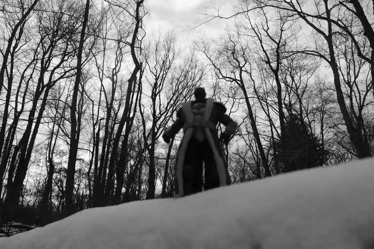 A black-and-white photo shows a small action figure, resembling a fantasy warrior or hero, standing on a snowy hill in a…