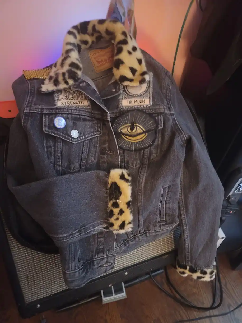 A dark-wash denim jacket with leopard-print faux fur collar and cuffs, adorned with patches including 'STRENGTH' and 'THE…