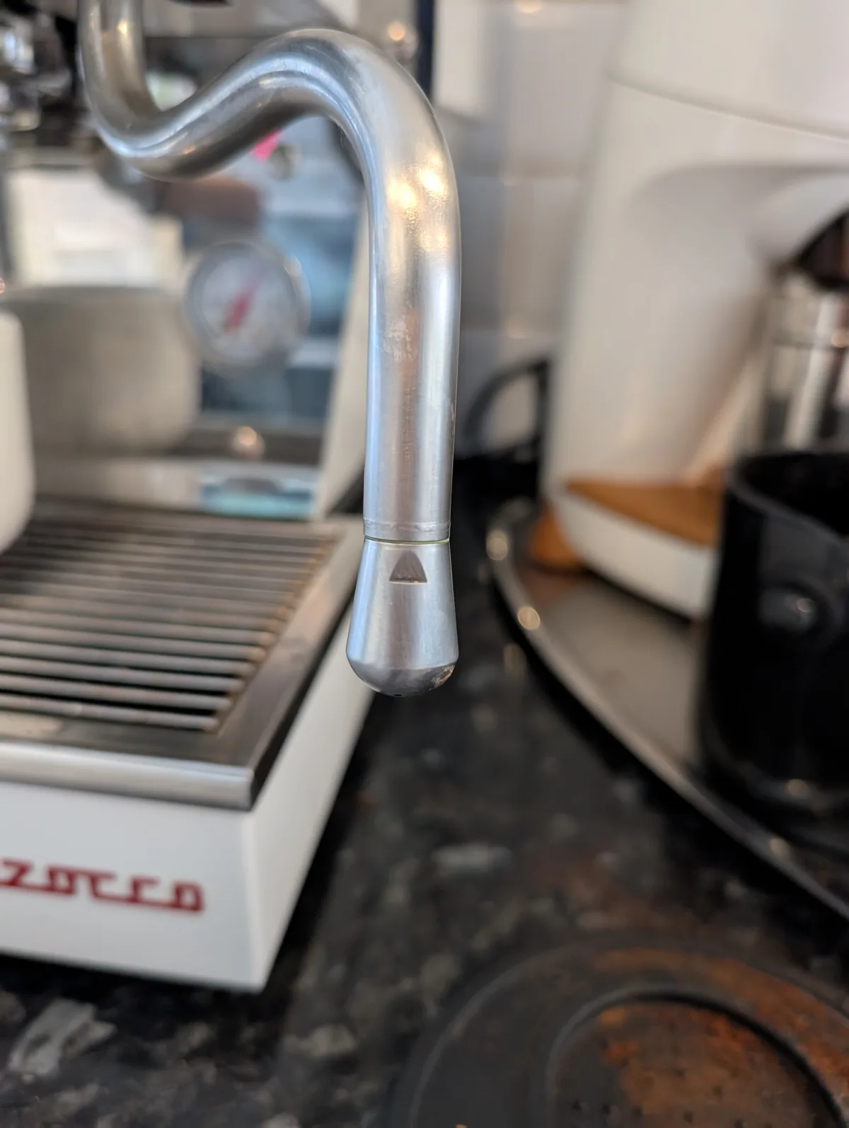 a close-up photo of a shiny, curved metal spout on an espresso machine, featuring a small triangular cutout at its tip. The…