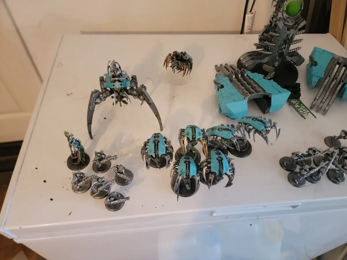 A collection of painted tabletop miniatures, mostly in a teal color scheme, are arranged on a white surface. The models…