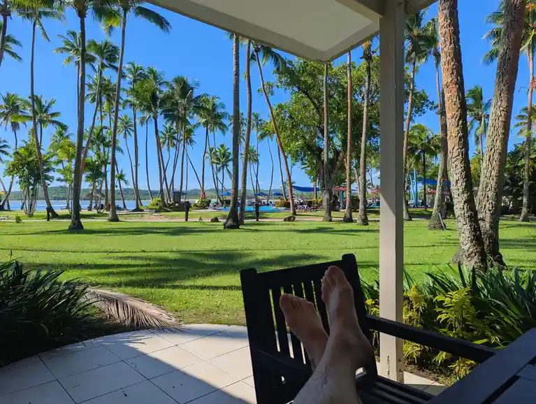 A first-person view from a covered porch shows bare feet resting on a black wooden chair, overlooking a lush green lawn…