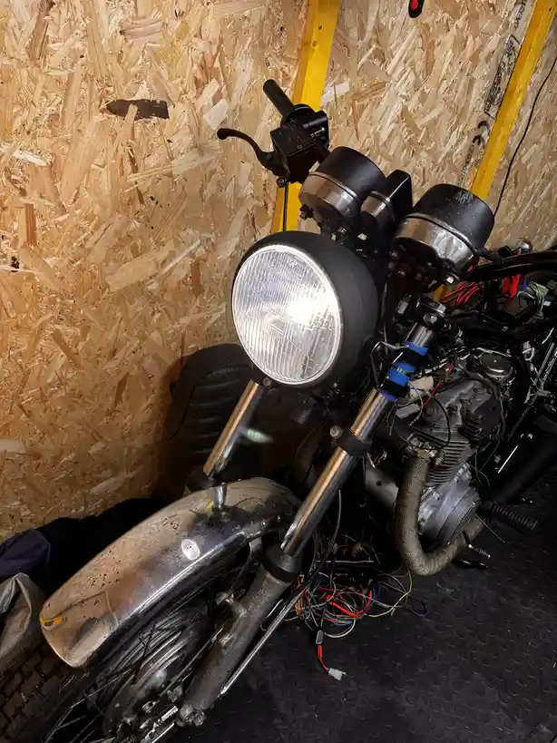 A partially disassembled motorcycle with exposed wiring and components sits in a garage with OSB walls, its headlight on…